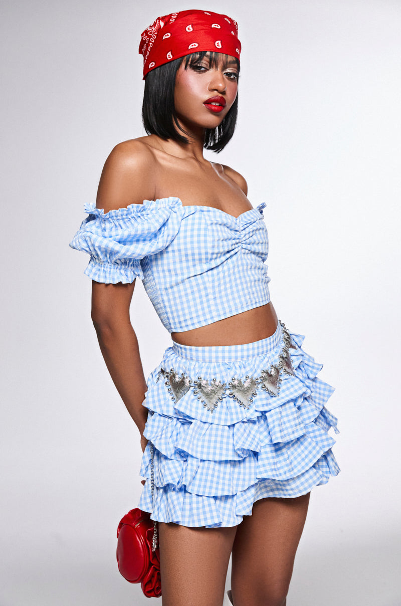 A person confidently poses in a blue and white gingham off-the-shoulder top with a ruffled skirt. They accessorize with a red bandana, red lipstick, holding a red bag, and the HOLD MY HEART METAL CHAIN BELT featuring heart pendants against a plain white backdrop.