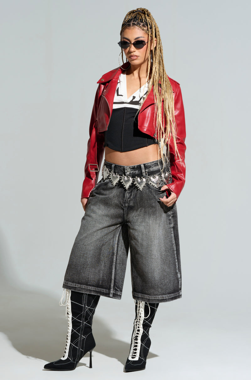 Against a gray background, a person with long braided hair and sunglasses strikes a confident pose wearing a red leather jacket, black and white corset top, oversized denim shorts, and knee-high black lace-up boots. The look is perfectly cinched at the waist with the edgy HOLD MY HEART METAL CHAIN BELT.
