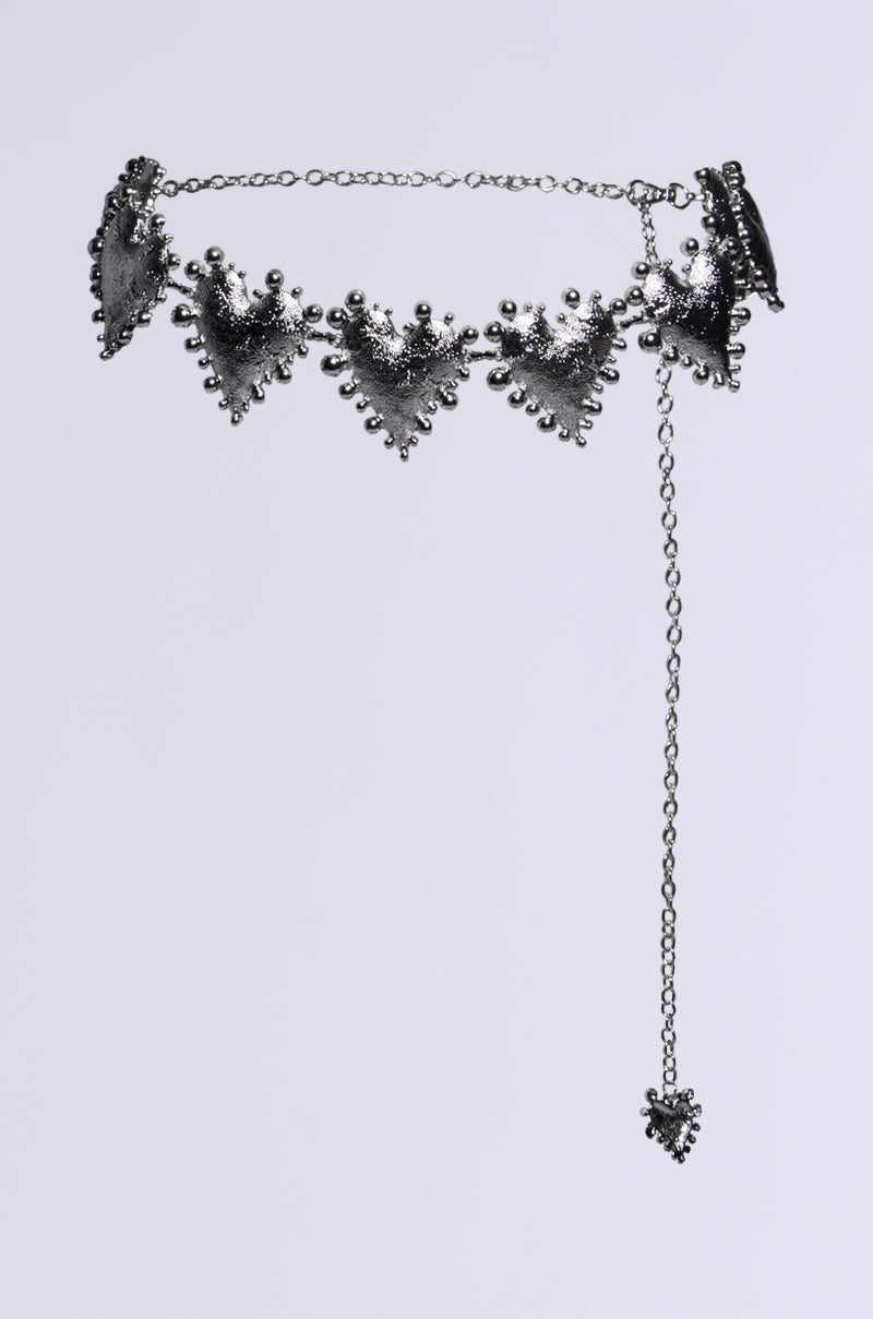 The HOLD MY HEART METAL CHAIN BELT is a silver accessory featuring a series of triangular, ornate pendants and an adjustable lobster clasp. It includes a long chain that extends down, concluding with a small heart pendant, all designed with intricate detailing that gives it a vintage appeal.