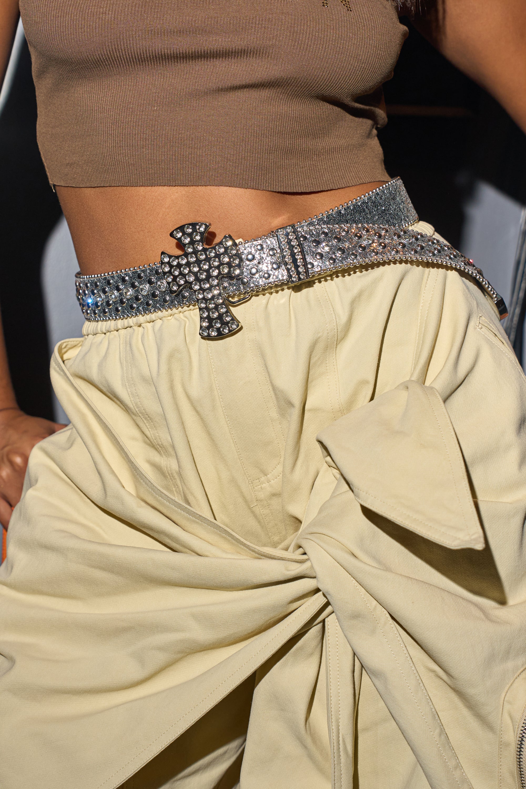 Wearing the IM BETTER BELT—a silver rhinestone belt with a cross-shaped western buckle—over high-waisted cream pants with a large side bow and a cropped brown top makes for the perfect festival accessory. 0