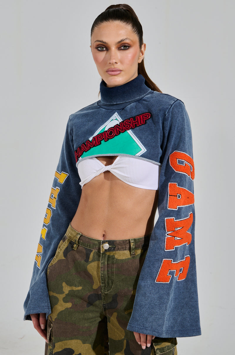 A woman confidently poses in the TRUE CHAMPION GRAPHIC CROPPED SWEATSHIRT, featuring bell sleeves and embroidered with "CHAMPIONSHIP" and "GAME." She pairs it with camo pants, sporting a high ponytail against a plain background.