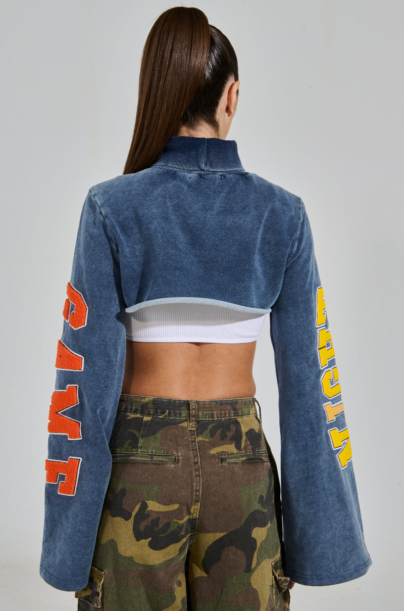 Rear view of a person with long hair wearing the TRUE CHAMPION GRAPHIC CROPPED SWEATSHIRT adorned with "GAME" and "KNIGHT" on the sleeves, revealing a white crop top underneath, paired with camouflage cargo pants.