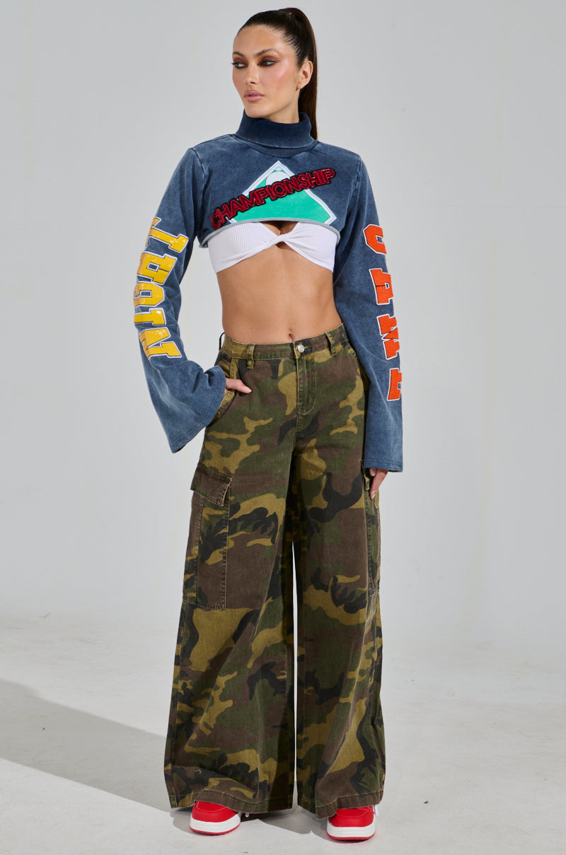 A person stands hands on hips wearing a blue TRUE CHAMPION GRAPHIC CROPPED SWEATSHIRT with colorful embroidered designs on sleeves and chest, paired with camouflage cargo pants and red sneakers against a plain light gray backdrop.