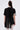 A person faces away, wearing the PEARL QUEEN OVERSIZED TEE—black, sheer, asymmetrical with center back pleats and rhinestone-trimmed short sleeves—paired with white lace-patterned leggings. The background is plain and light-colored.