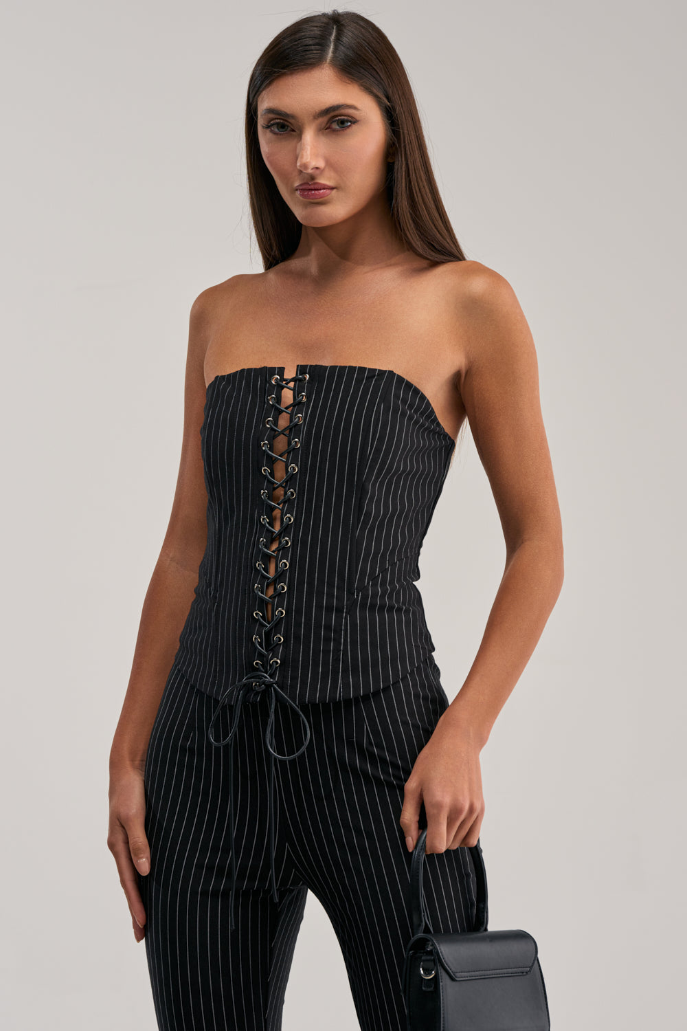 A woman in the LIVI STRETCHY PINSTRIPE CORSET with matching pinstriped pants holds a small black handbag, standing against a plain light gray background.