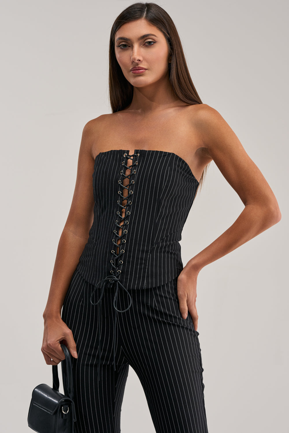 A woman wears the LIVI STRETCHY PINSTRIPE CORSET with a strapless neckline and front lace-up detail, paired with matching pinstripe pants. She stands confidently, one hand on her hip and holding a small black handbag.