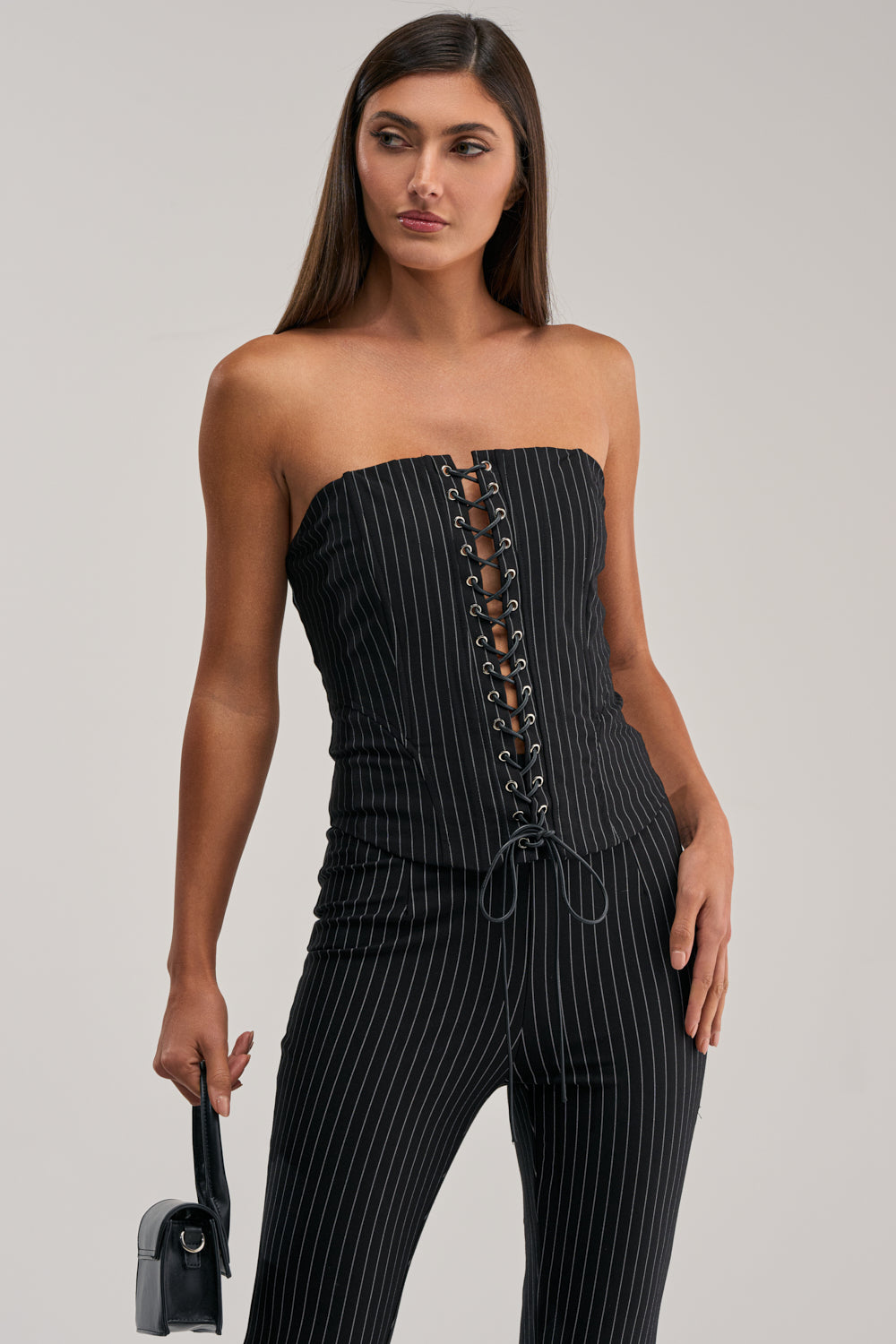 A woman stands holding a small black handbag, wearing the LIVI STRETCHY PINSTRIPE CORSET—a strapless black jumpsuit with a pinstripe corset top and lace-up front—looking slightly to the side against a plain background.