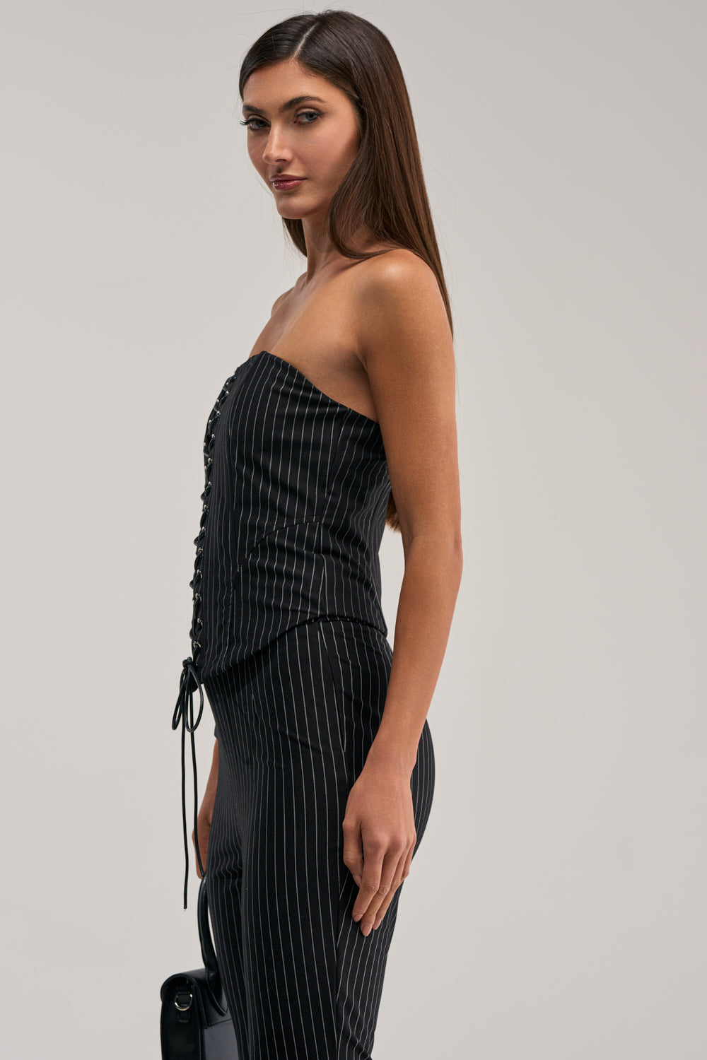 A woman with long straight brown hair poses sideways against a light backdrop wearing the LIVI STRETCHY PINSTRIPE CORSET and matching pants, holding a black bag. The corset features a strapless neckline and pinstripe design.