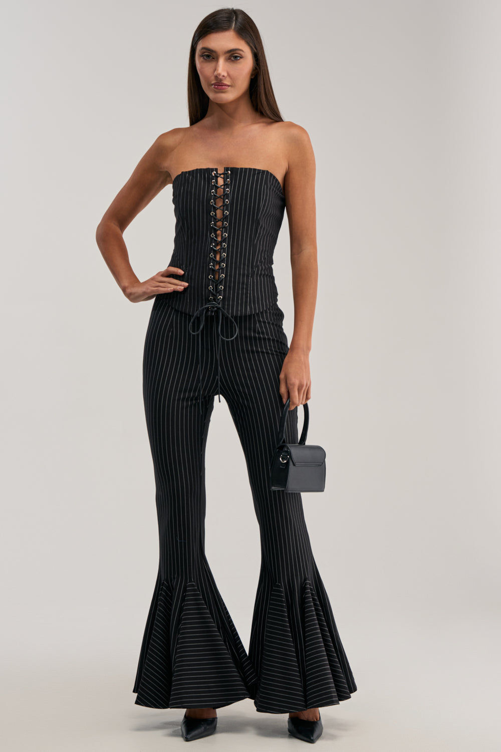 A woman stands against a light background wearing LIVI STRETCHY PINSTRIPE FLARE LEGGINGS, paired with a strapless black top, black heels, and holding a small black handbag. The leggings feature pinstripes and flared bell-bottom legs.