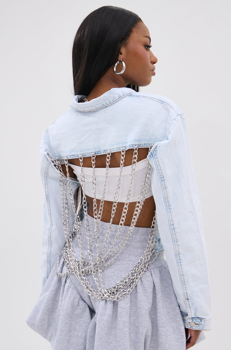 A woman with long dark hair wears the PARTY IN THE BACK DENIM JACKET—an acid-washed denim piece with an open back and silver-tone chains. She pairs it with large hoop earrings, a white top, and gray high-waisted pants.