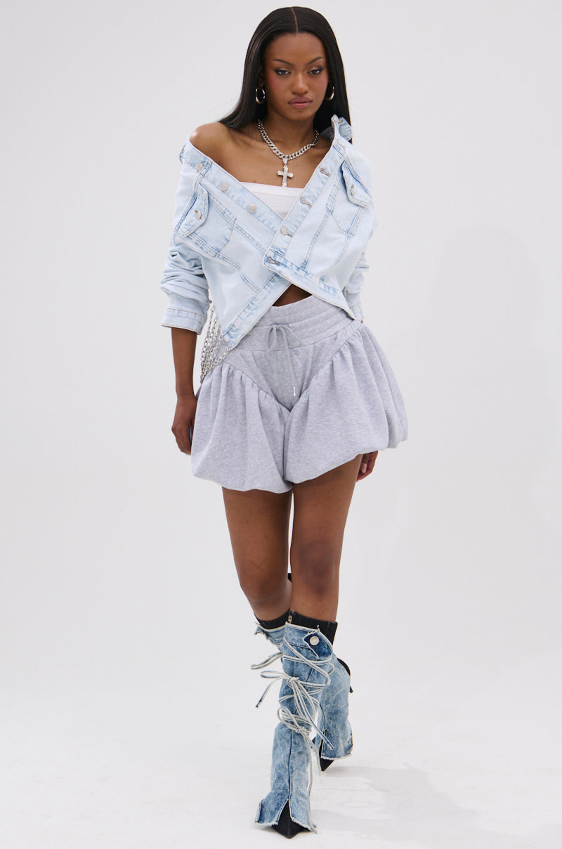 A woman walks confidently in the PARTY IN THE BACK DENIM JACKET—light blue, acid-washed, off-shoulder style—with a grey bubble mini skirt, top, layered silver-tone chains, denim lace-up leg warmers over black shoes on a white background.