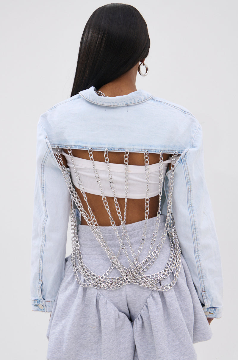 A person with long dark hair wears the PARTY IN THE BACK DENIM JACKET, featuring an acid wash, open back with draped silver-tone chains over a white bandeau top, paired with high-waisted light grey voluminous pants.