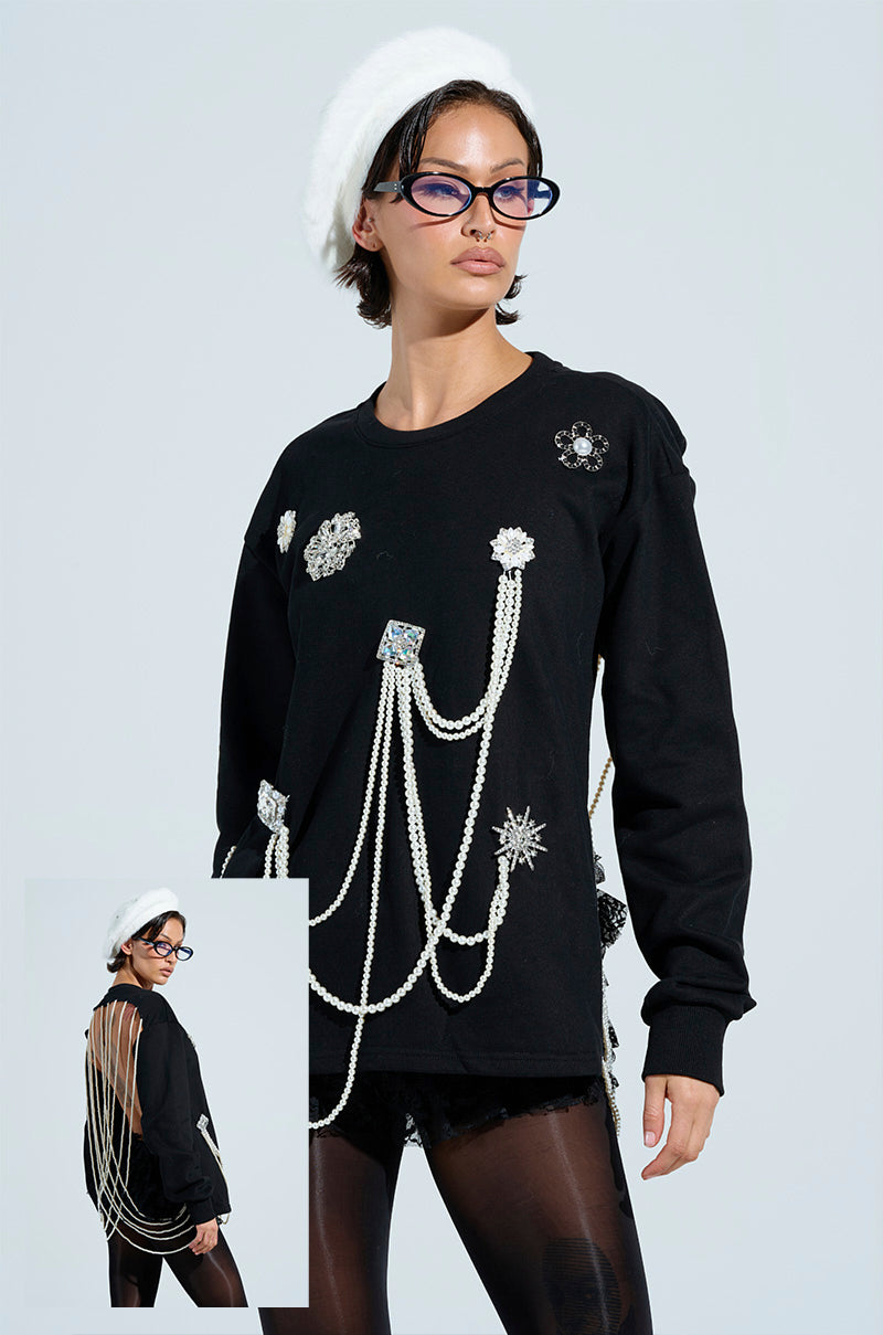A person in the IT'S COMPLICATED PEARL TOP, a black sweatshirt with white pearl accents and silver motifs, paired with glasses and a white beret. An inset reveals chain details and a flower brooch on the back.