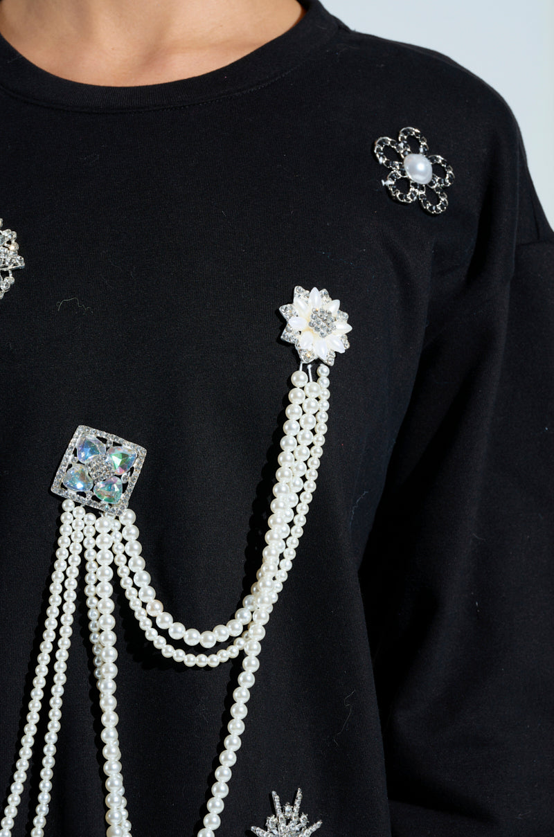A close-up of the IT'S COMPLICATED PEARL TOP, a black sweater adorned with pearl beads and rhinestone brooches in floral and geometric shapes, highlighting its elegant decorative detailing.