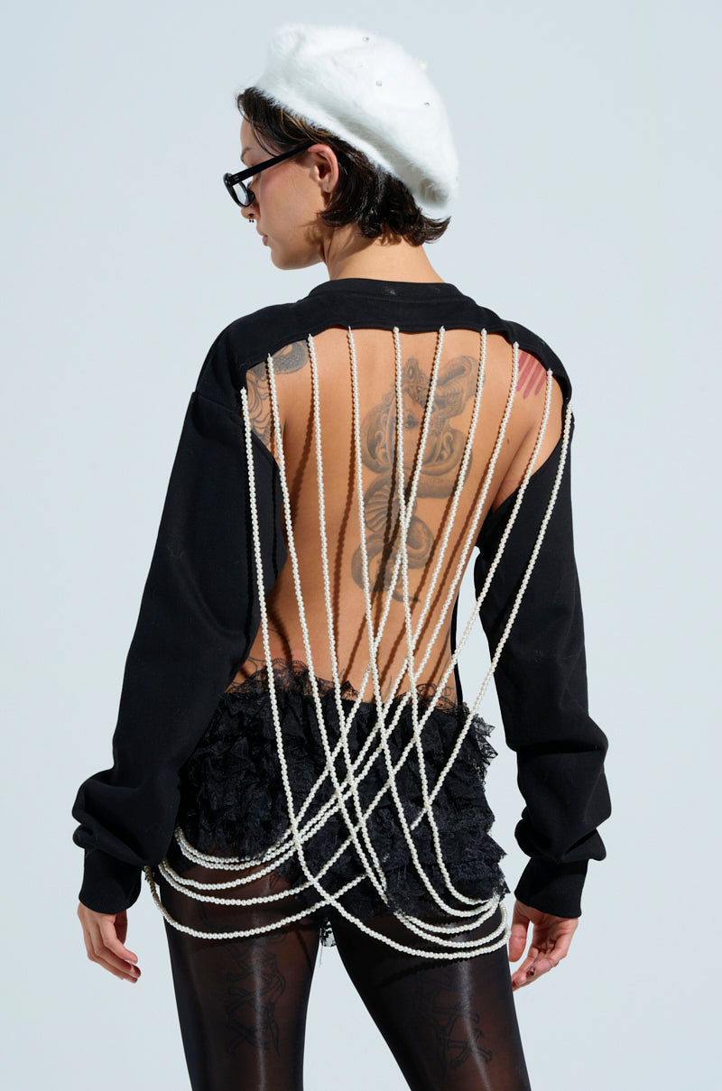 A person wears the IT'S COMPLICATED PEARL TOP—a black open-back outfit with crisscrossing white strings and beaded pearl accents. They have back tattoos, a white beret with flower brooches, and black glasses.
