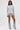 A woman with straight hair, hoop earrings, and layered necklaces stands against a white background wearing the OPEN BACK CHAIN DETAIL SWEATSHIRT IN GREY, matching shorts, and loose gray leg warmers over heeled shoes.