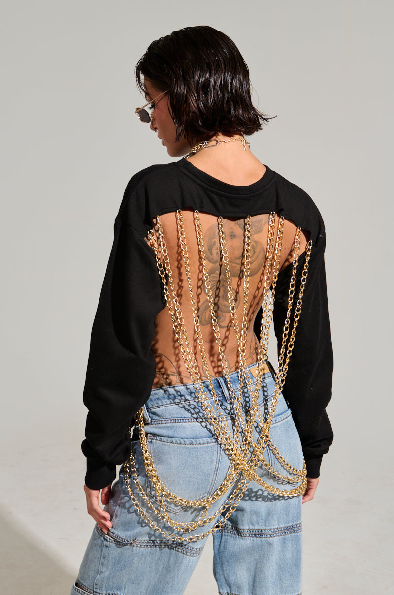 A person wearing sunglasses, an OPEN BACK CHAIN DETAIL SWEATSHIRT styled like a chic black top, and jeans featuring elegant gold chain accents draped across their back and waist. The background is simple and minimal, creating an effortlessly casual look.