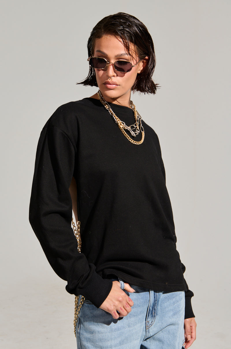 A person with short, dark hair sports a casual look with sunglasses and an OPEN BACK CHAIN DETAIL SWEATSHIRT in black. They pair it with light blue jeans and gaze off to the side against a plain background.