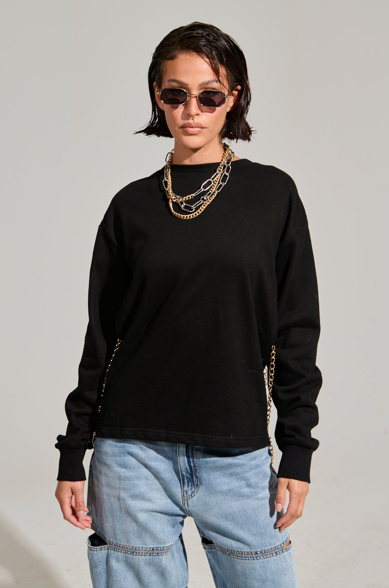 A person with short dark hair is wearing sunglasses, an OPEN BACK CHAIN DETAIL SWEATSHIRT, and loose blue jeans. Their casual look is completed with a chain necklace. They are standing against a plain gray background, looking confidently at the camera.