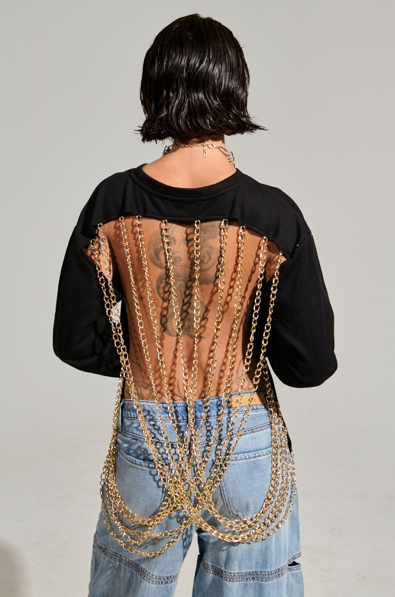 A person with short dark hair is facing away, wearing the OPEN BACK CHAIN DETAIL SWEATSHIRT, which features gold chains elegantly draped in cascading layers down the open back. They pair it with faded blue jeans with rips and have a visible tattoo on their upper back, creating a chic yet casual look.