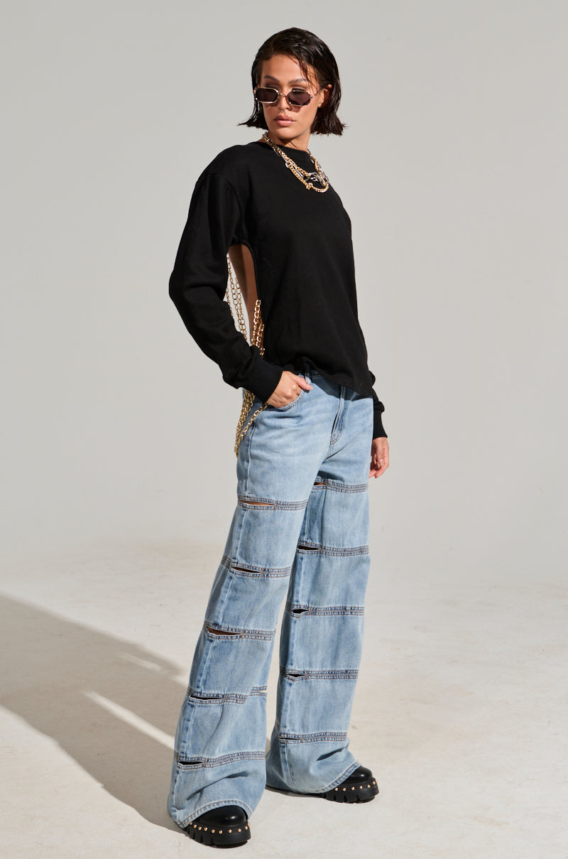 Wearing an OPEN BACK CHAIN DETAIL SWEATSHIRT in black with side cutouts and paired with light blue wide-leg jeans featuring multiple horizontal seams, a person showcases their casual style. Sporting short dark hair, sunglasses, and chunky black shoes, they pose confidently against a plain backdrop.