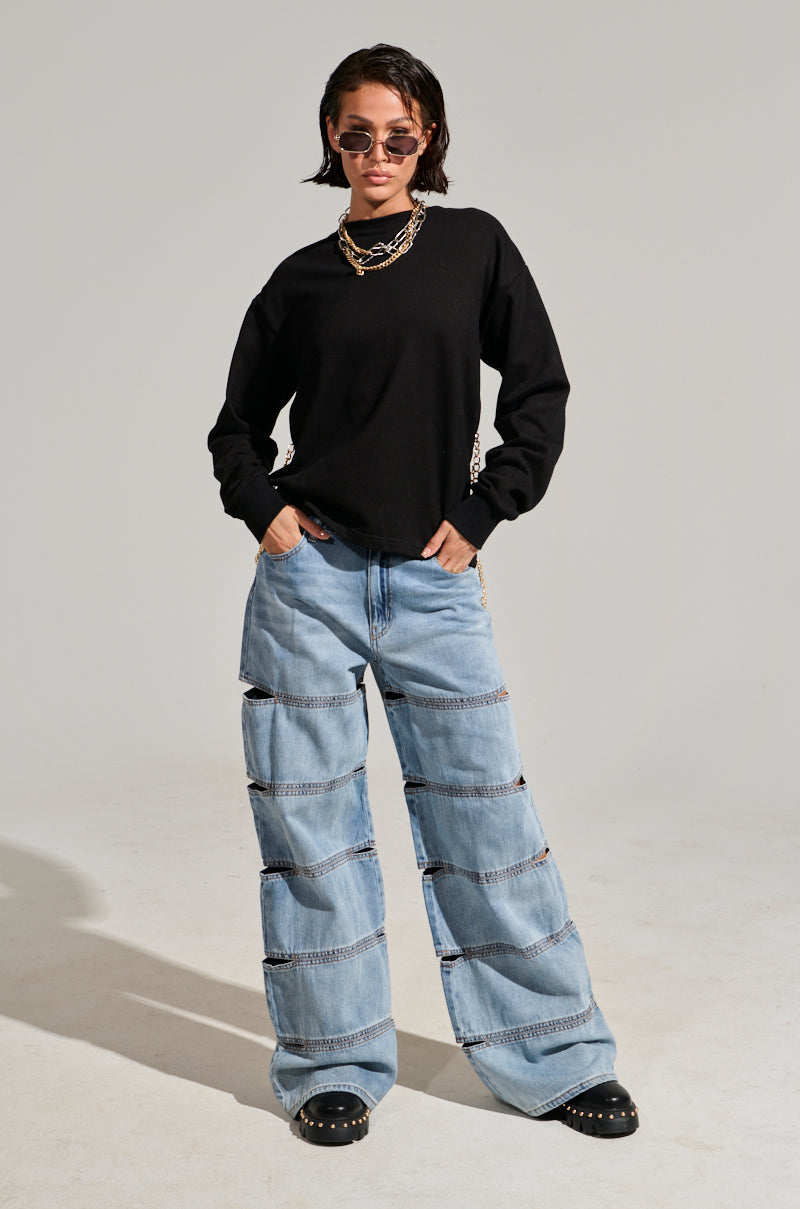 A person stands confidently in an Open Back Chain Detail Sweatshirt, layered gold chain detail necklaces, and oversized light blue jeans with horizontal stripe patterns. They accessorize with round sunglasses and black studded boots for a chic yet casual look.
