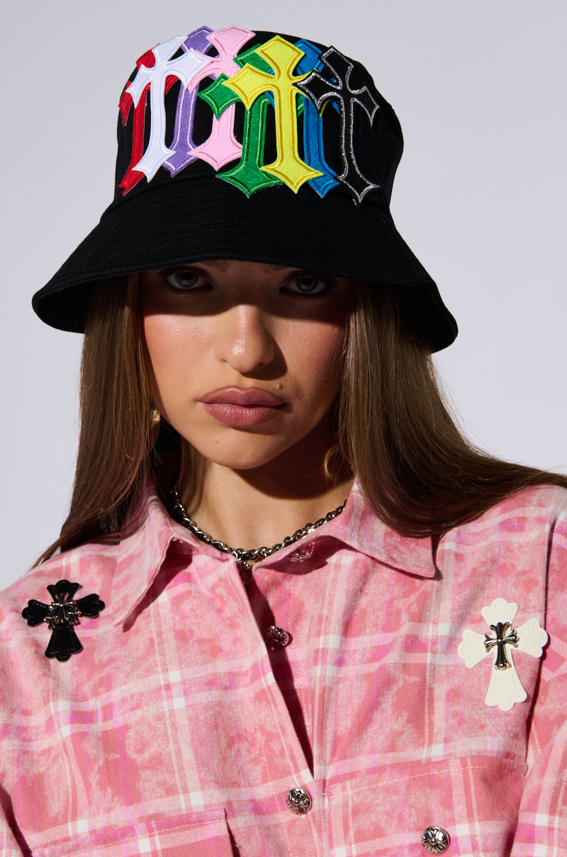 A woman with long brown hair wears a CROSSED PATCH BUCKET HAT with multicolored embroidered letters and a pink plaid shirt adorned with cross-shaped brooches. She has a serious expression and a chain necklace. The background is plain and light-colored, highlighting her distinct style featuring embroidered cross patches. 0