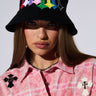 A woman with long brown hair wears a CROSSED PATCH BUCKET HAT with multicolored embroidered letters and a pink plaid shirt adorned with cross-shaped brooches. She has a serious expression and a chain necklace. The background is plain and light-colored, highlighting her distinct style featuring embroidered cross patches.