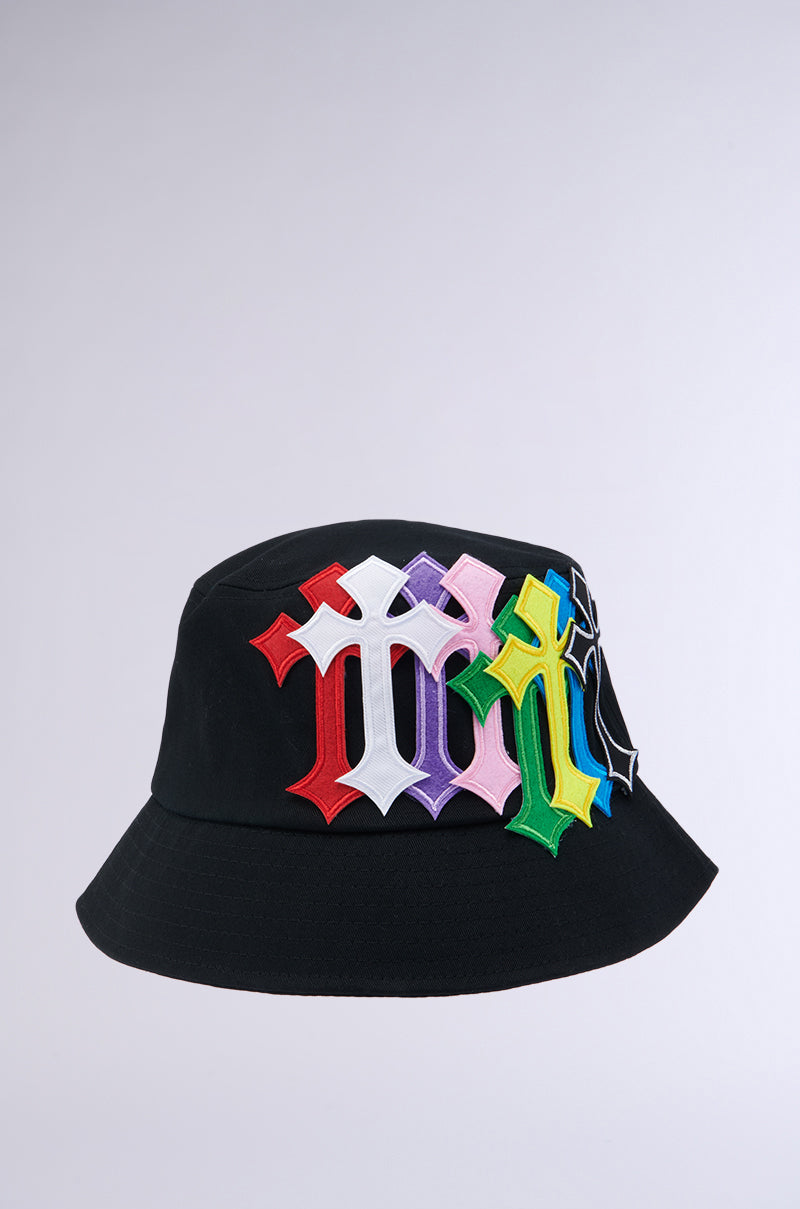 The CROSSED PATCH BUCKET HAT is a black cotton base hat adorned with colorful, overlapping Gothic-style letter "M" patches in shades of red, white, purple, pink, green, yellow, and blue. These patches are meticulously arranged on a plain light gray background. 1