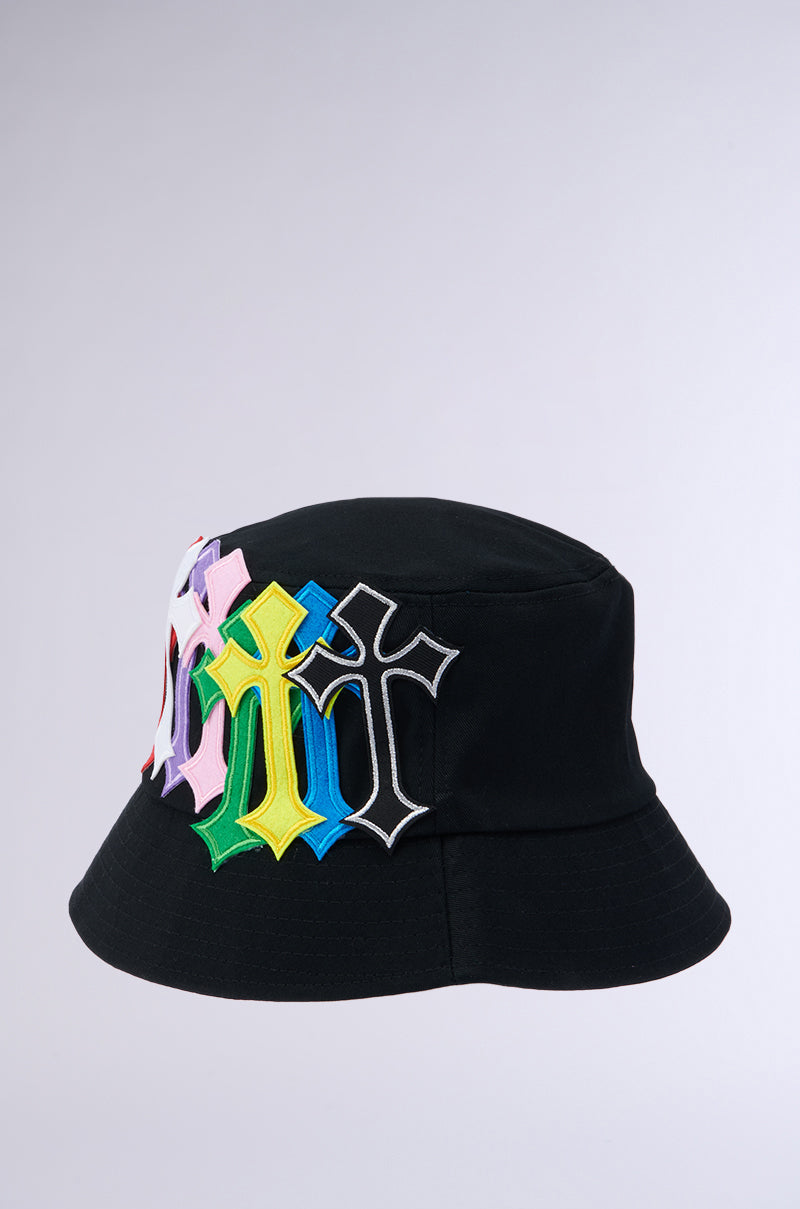 The CROSSED PATCH BUCKET HAT features a black cotton base accentuated by embroidered cross patches in shades of pink, yellow, blue, white, and black, creating a vibrant contrast against a plain light grey background. 3