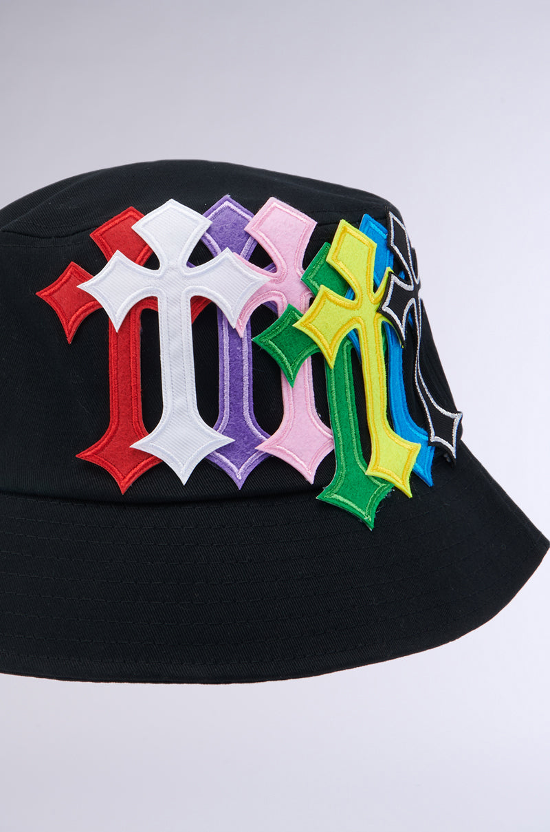 The CROSSED PATCH BUCKET HAT from the AKIRA Label features a cotton base adorned with multiple Gothic-style letter "M" patches in an array of vibrant colors, including red, white, purple, pink, green, yellow, blue, and black. These embroidered cross patches are prominently overlapping on the hat's front side against a plain light-colored background. 4