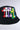 The CROSSED PATCH BUCKET HAT from the AKIRA Label features a cotton base adorned with multiple Gothic-style letter "M" patches in an array of vibrant colors, including red, white, purple, pink, green, yellow, blue, and black. These embroidered cross patches are prominently overlapping on the hat's front side against a plain light-colored background.