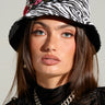 A person with long brown hair is wearing the ROSES ARE RED ZEBRA BUCKET HAT, featuring red rose embroidery. They are dressed in a black sheer top and a chunky silver chain necklace. Their vibrant look is tied together by the floral embroidered detail on the hat, set against a plain light grey background.