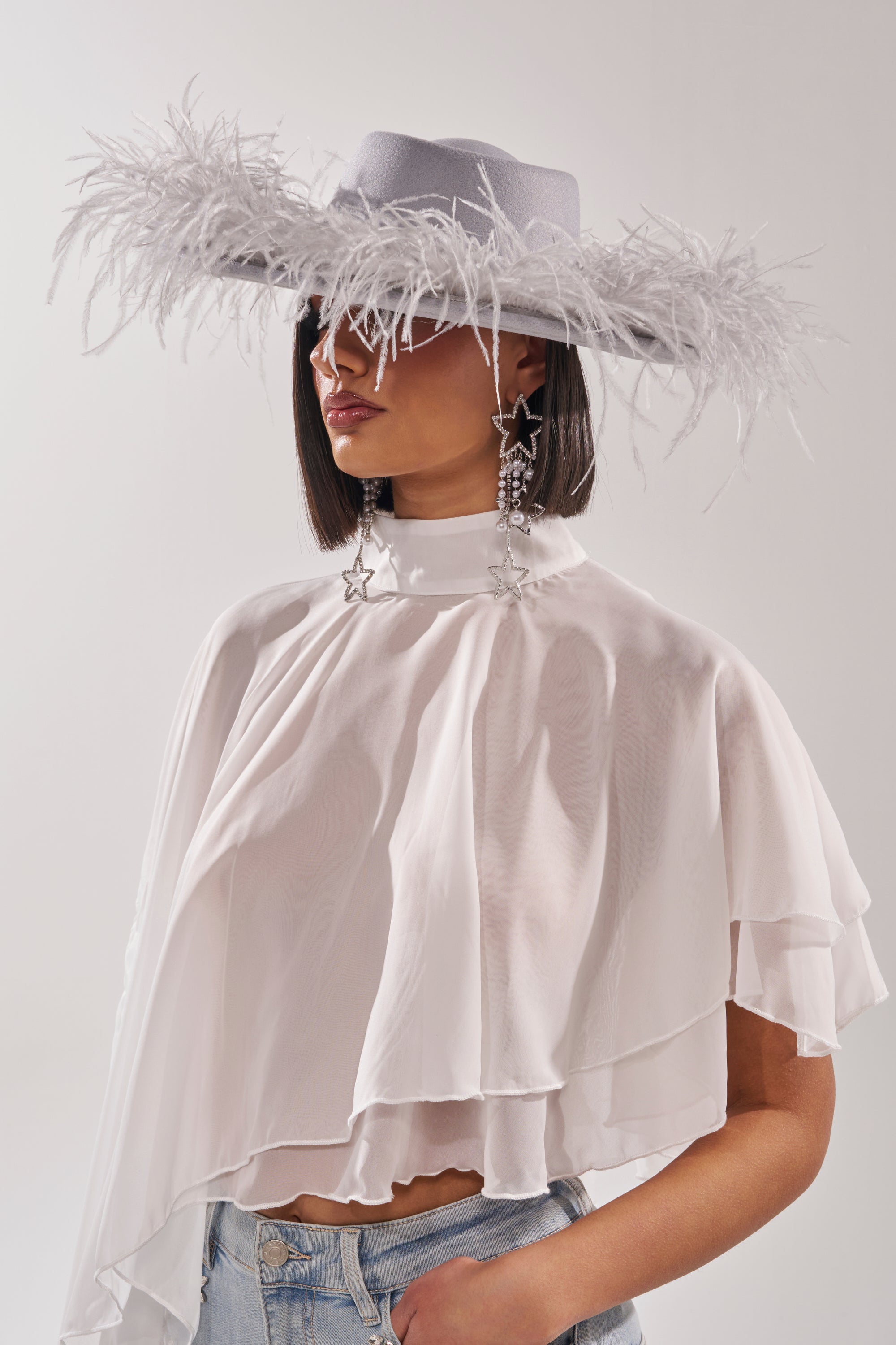 A woman wears the FEEL IT FEATHER BRIM HAT—blue with pearl trim—paired with star-shaped earrings, a sheer white layered top, and light blue jeans. She stands before a pale background, her eyes concealed by this standout festival piece. 0