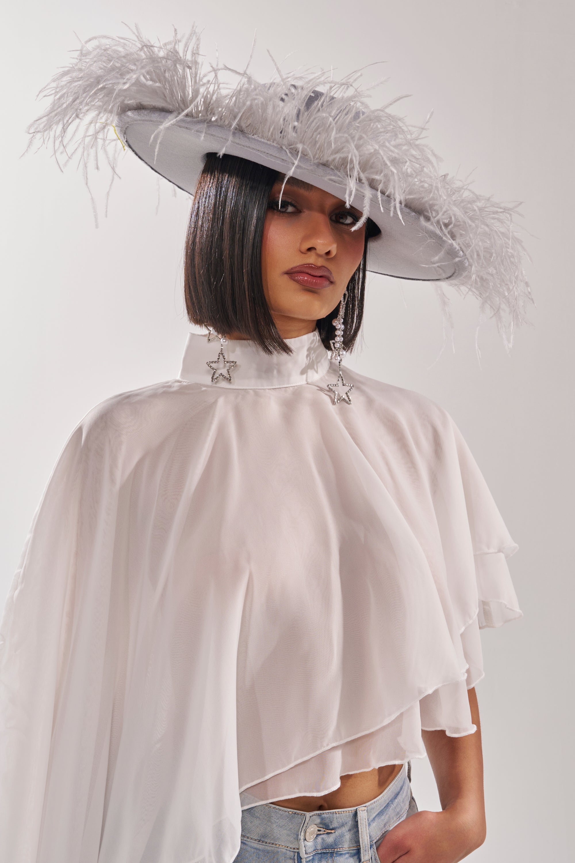 A person wearing the FEEL IT FEATHER BRIM HAT in large blue, paired with star-shaped earrings and a flowing white cape-style top, poses against a plain background with straight dark hair and a neutral expression. 1