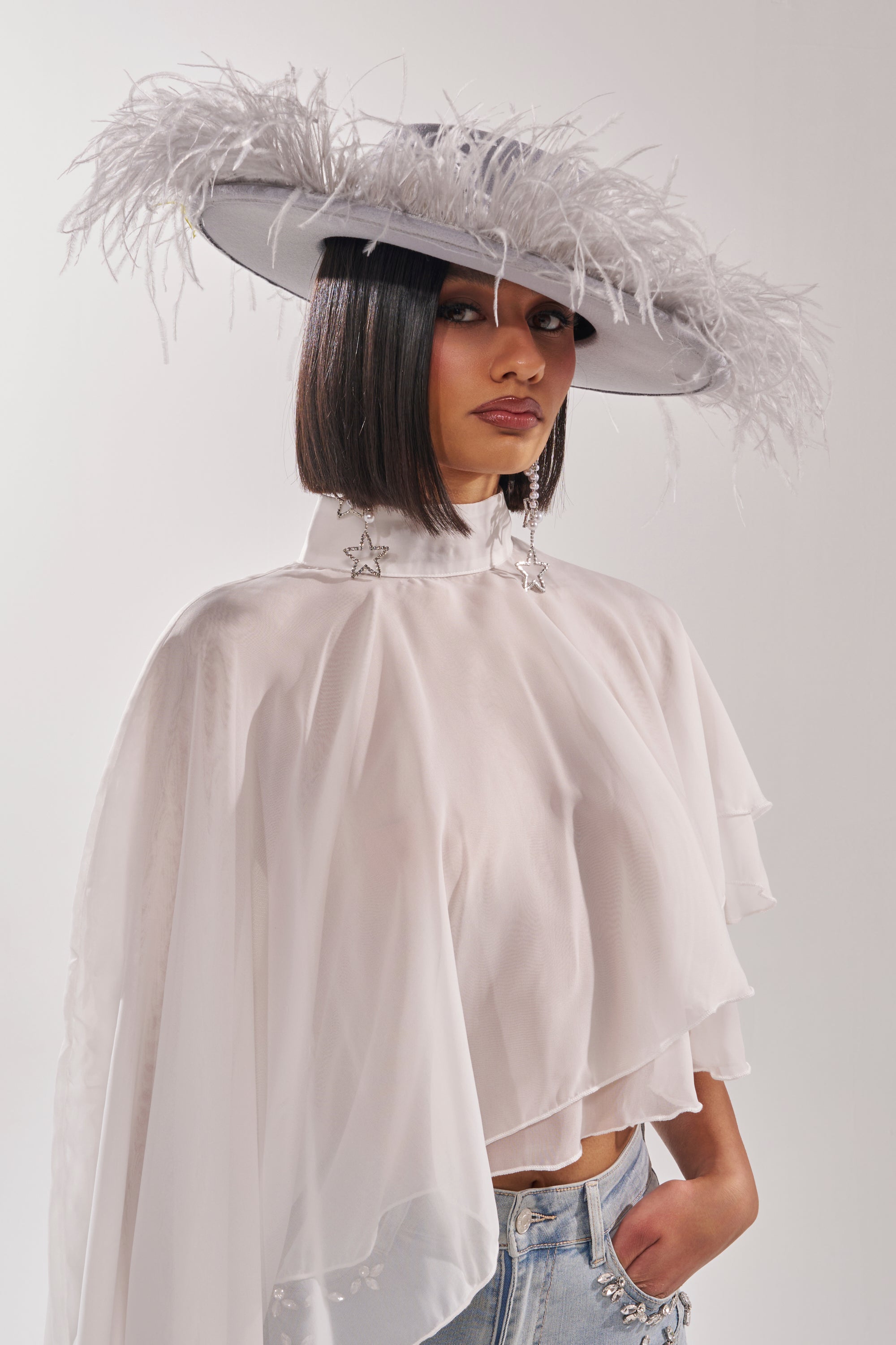 A person with a sleek bob wears the FEEL IT FEATHER BRIM HAT, a wide-brimmed blue feather cowboy hat, styled with a sheer white blouse, star earrings, and light blue jeans against a plain light background. 2