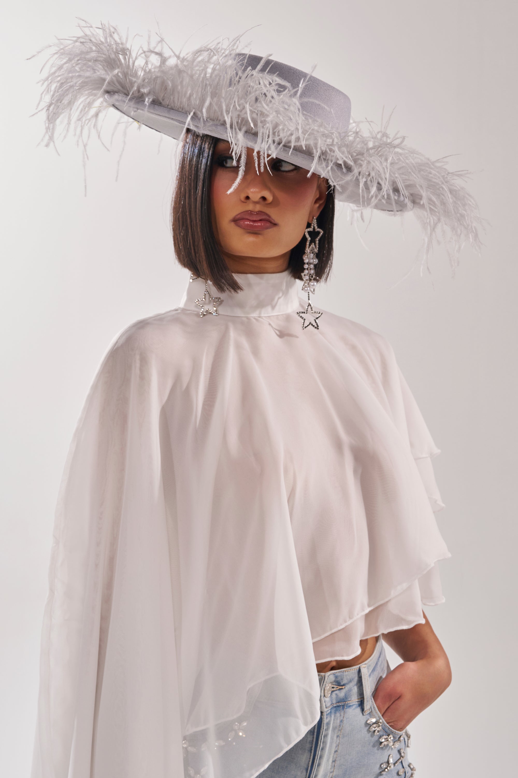 Wearing the FEEL IT FEATHER BRIM HAT in blue with pearl trim, a model poses confidently in a sheer white cape top, high-waisted jeans, and star earrings against a plain light background. 3
