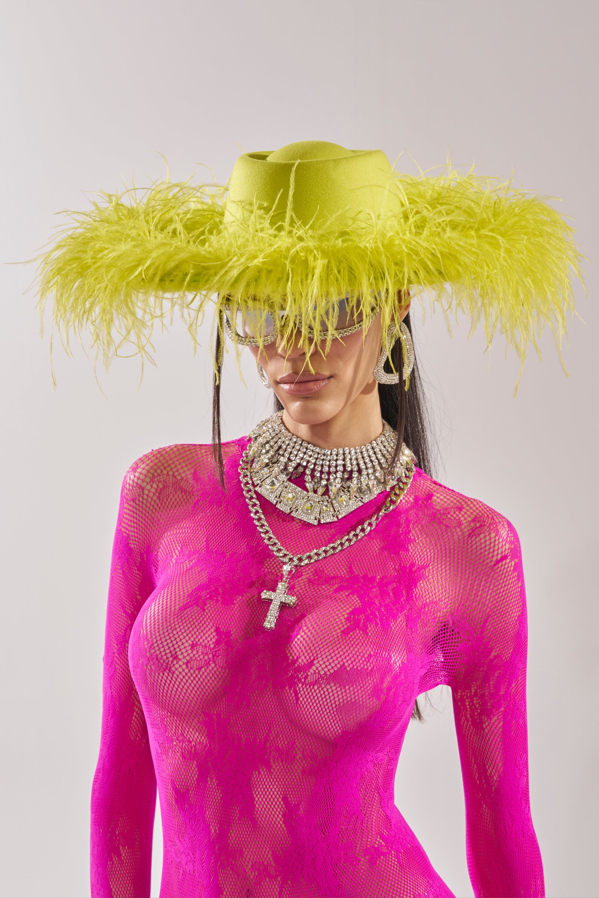 A person wears a sheer, bright pink lace outfit with layered silver necklaces and the FEEL IT FEATHER BRIM HAT IN GREEN. They accessorize with oversized sunglasses and statement earrings against a plain background. 1