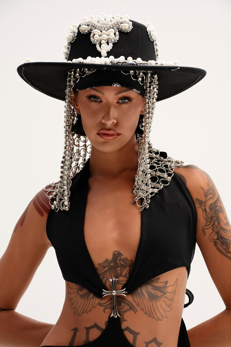 A tattooed and pierced individual dons the PEARL PLEASE EMBELLISHED BRIM HAT, lavishly adorned with pearls and chains. They wear a sleeveless black top with metallic accents, standing out against a plain background, exuding eclectic style. 4