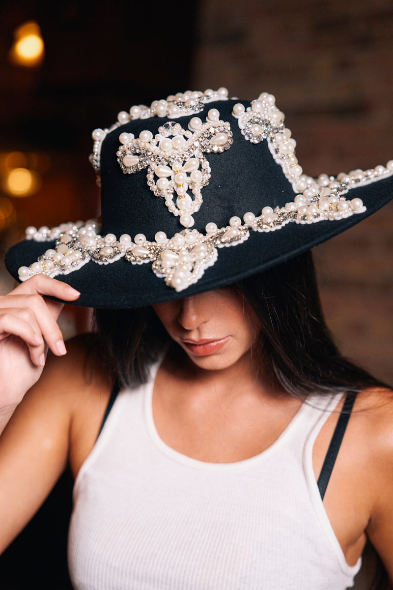 A woman in a white tank top tilts the PEARL PLEASE EMBELLISHED BRIM HAT, detailed with pearls and jewels, partially covering her face against a softly blurred background. 1