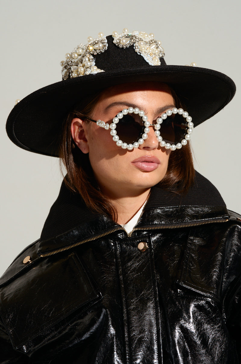 A person wearing the PEARL PLEASE EMBELLISHED BRIM HAT and a pair of large, round sunglasses adorned with pearls. The individual is dressed in a shiny black jacket adorned with rhinestone details and appears to be looking forward. 8