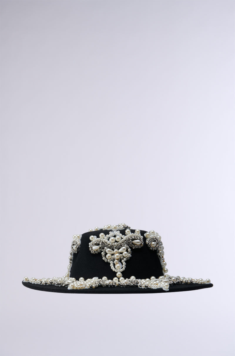 The PEARL PLEASE EMBELLISHED BRIM HAT is a dark-colored wide-brim hat adorned with ornate pearl and rhinestone details, showcasing intricate designs on the crown and brim. The plain and indistinct background highlights the detailed decorative elements of this exquisite piece.