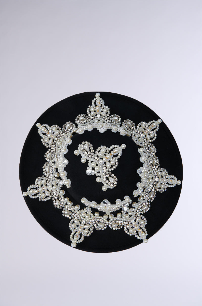 The PEARL PLEASE EMBELLISHED BRIM HAT is a black velvet hat adorned with intricate white and silver beaded embellishments that form circular and floral patterns. This festive hat features a central cluster of beads and an embellished brim that extends towards the edges, creating an elegant and eye-catching design. 11