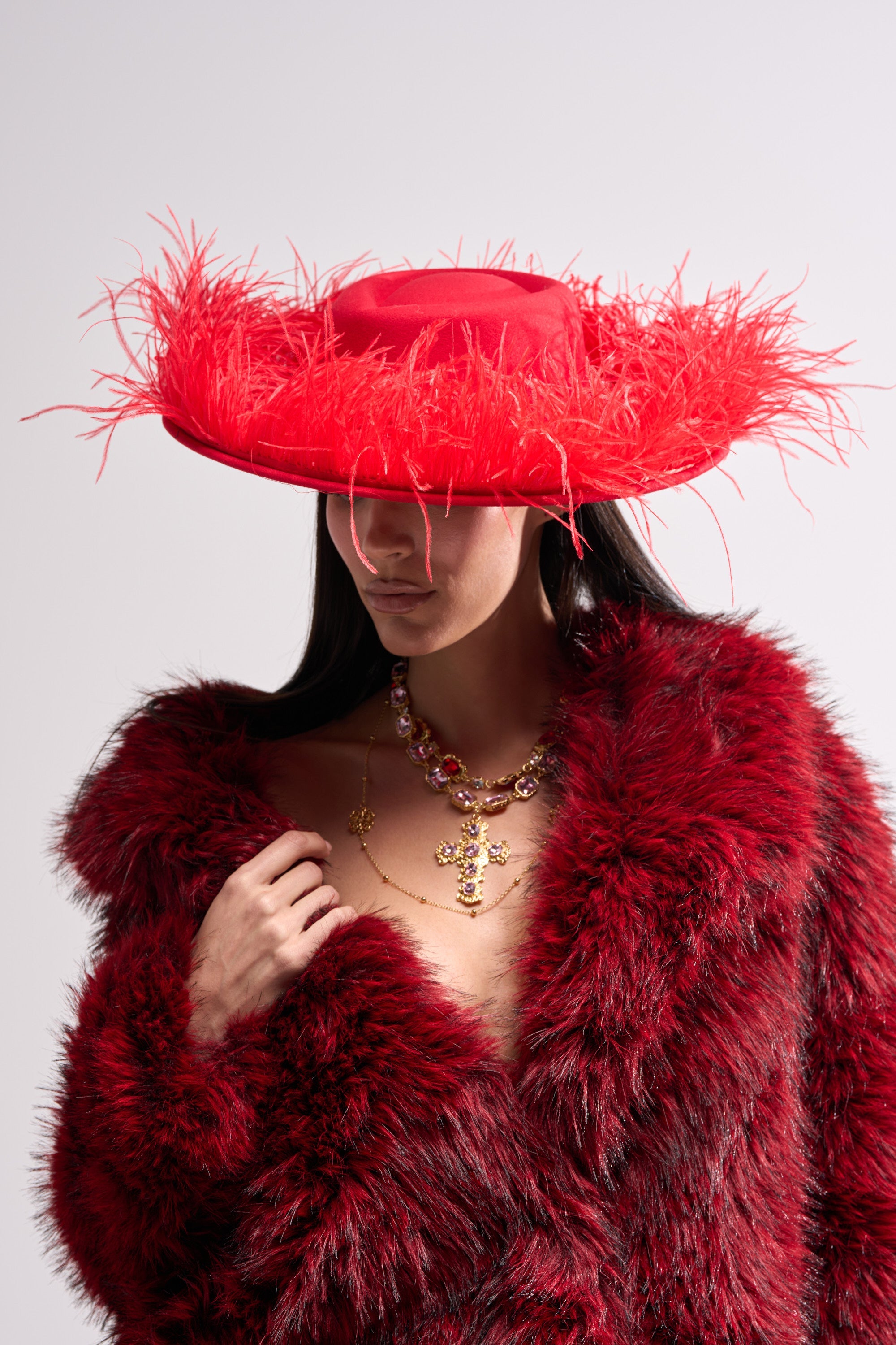 A woman poses against a plain background in a bright red LETS FLIRT FEATHER BRIM HAT and a red faux fur coat; her face is partly hidden by the hat’s wide brim, and she finishes the look with layered gold necklaces. 0