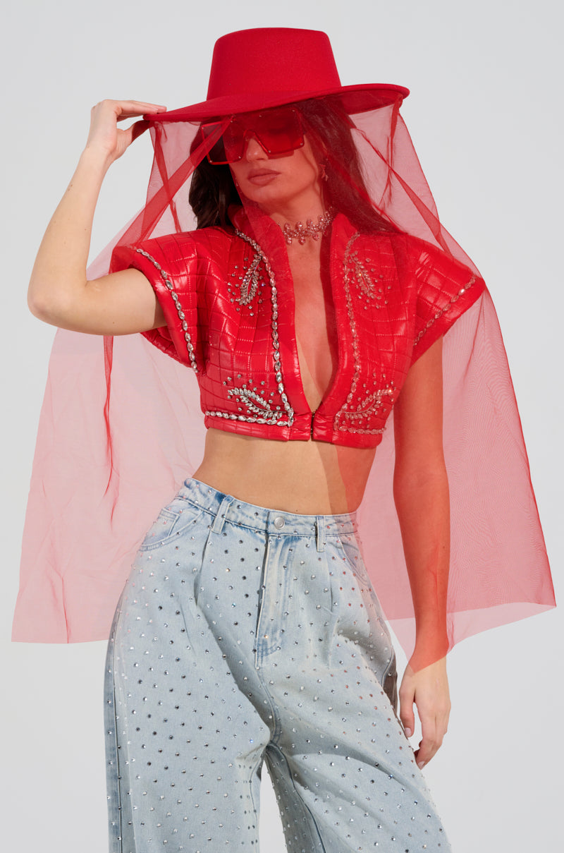 A person poses against a plain background, wearing a TYPICAL ELEGANCE MESH OVERLAY BRIM HAT in red, which features a tonal mesh veil. They pair it with a red beaded top and silver denim jeans embellished with rhinestones, completing the look with red sunglasses and a necklace. 0