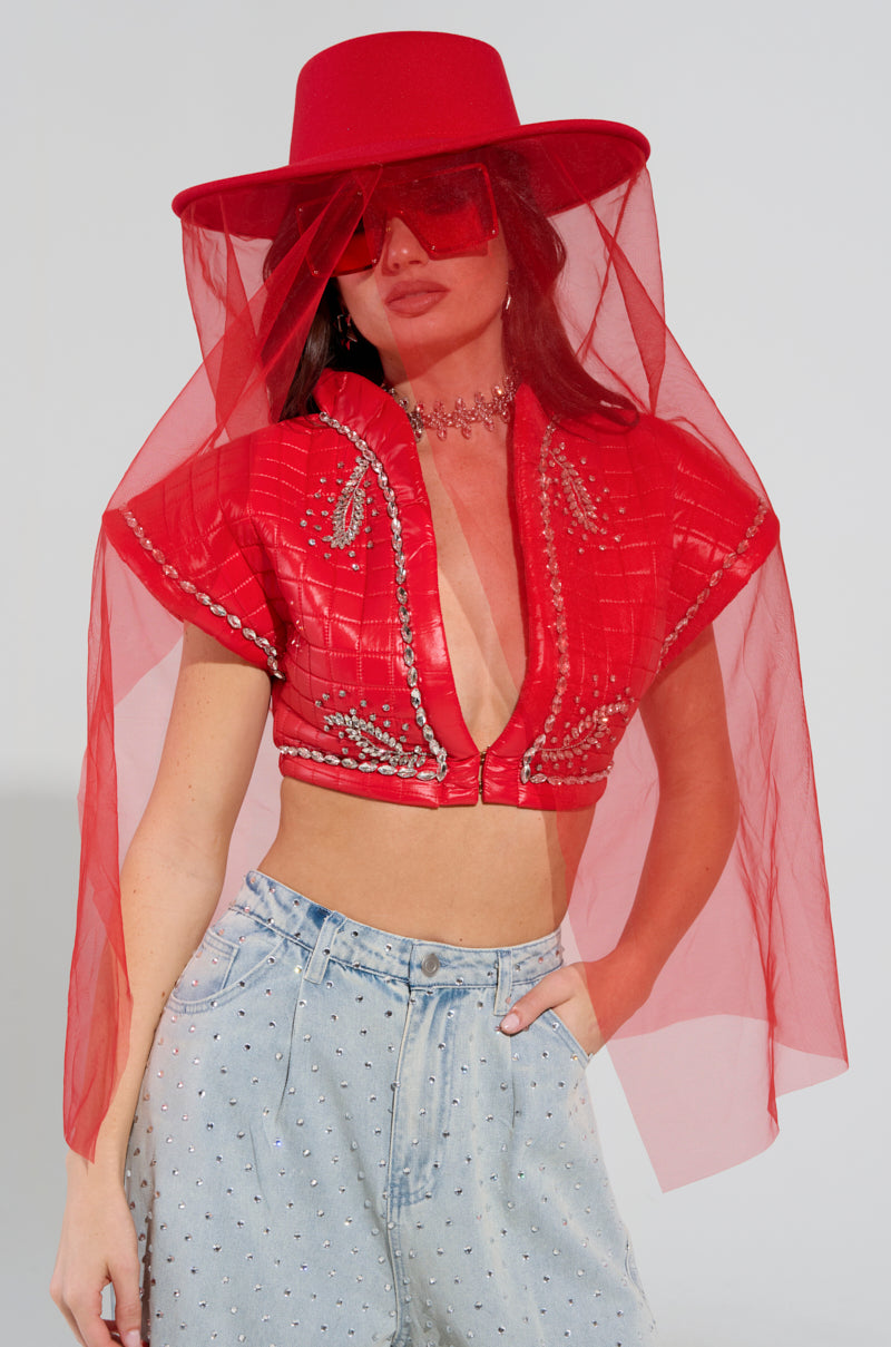Dressed in a striking red ensemble, a woman showcases the TYPICAL ELEGANCE MESH OVERLAY BRIM HAT by AKIRA Label, complemented by red sunglasses and a cropped top adorned with intricate embellishments. Her look is completed with high-waisted light denim jeans featuring small stud details. 1