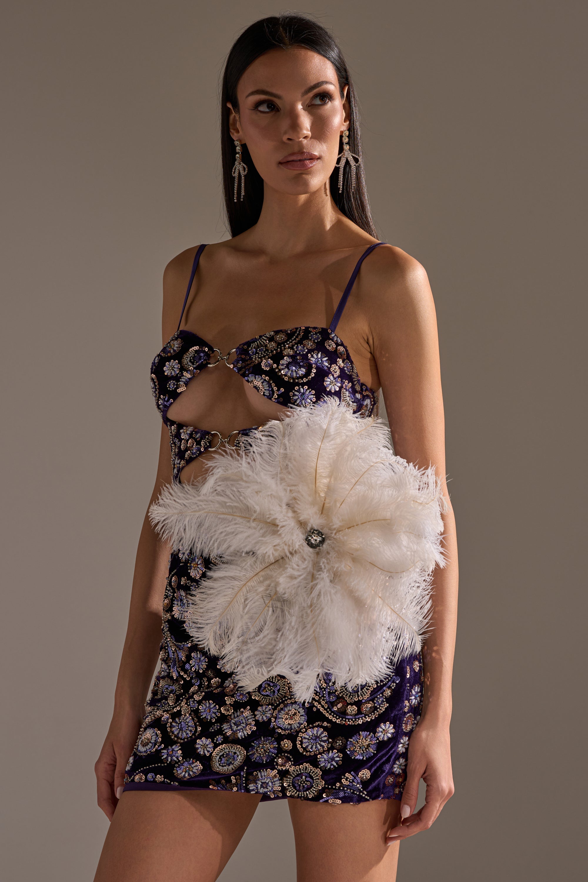 A woman in a dark embellished mini dress with cut-out details styles the TREAT YOURSELF FEATHER BROOCH, featuring a large white feather and gemstone center, while standing against a neutral background. 1