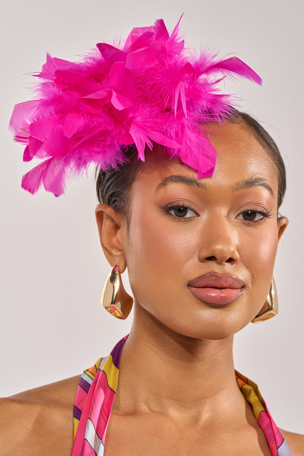 A woman with smooth skin, bold gold earrings, a colorful halter top, and a bright pink feathered fascinator finishes her subtle makeup with the AT BRUNCH FEATHER BROOCH. The background is plain and light. 0