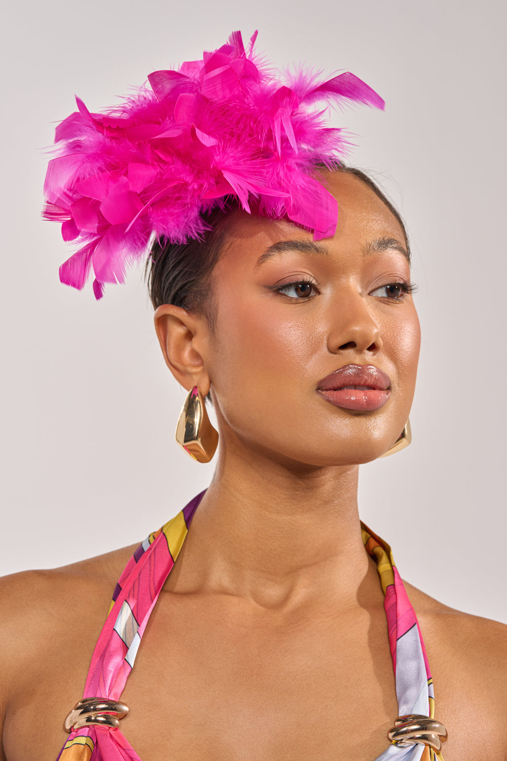 A woman poses confidently against a neutral background, wearing gold hoop earrings, a colorful halter top, and the AT BRUNCH FEATHER BROOCH—a bold fuchsia accessory that adds a striking touch to her look. 1