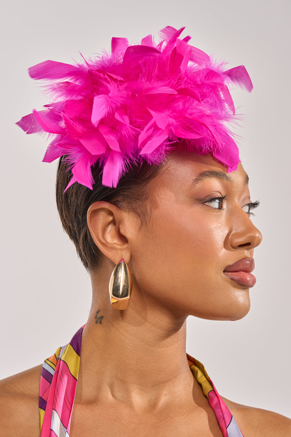 A person with short hair wears a bright pink feathered headpiece, gold geometric earrings, and a colorful top, styled with the AT BRUNCH FEATHER BROOCH in fuchsia, looking to the right against a neutral background. 2
